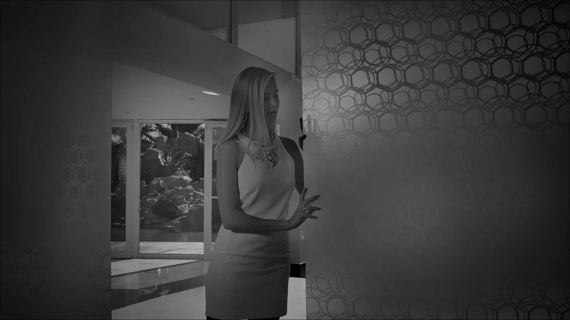 A woman stands beside a patterned wall, with a view of a lush garden.