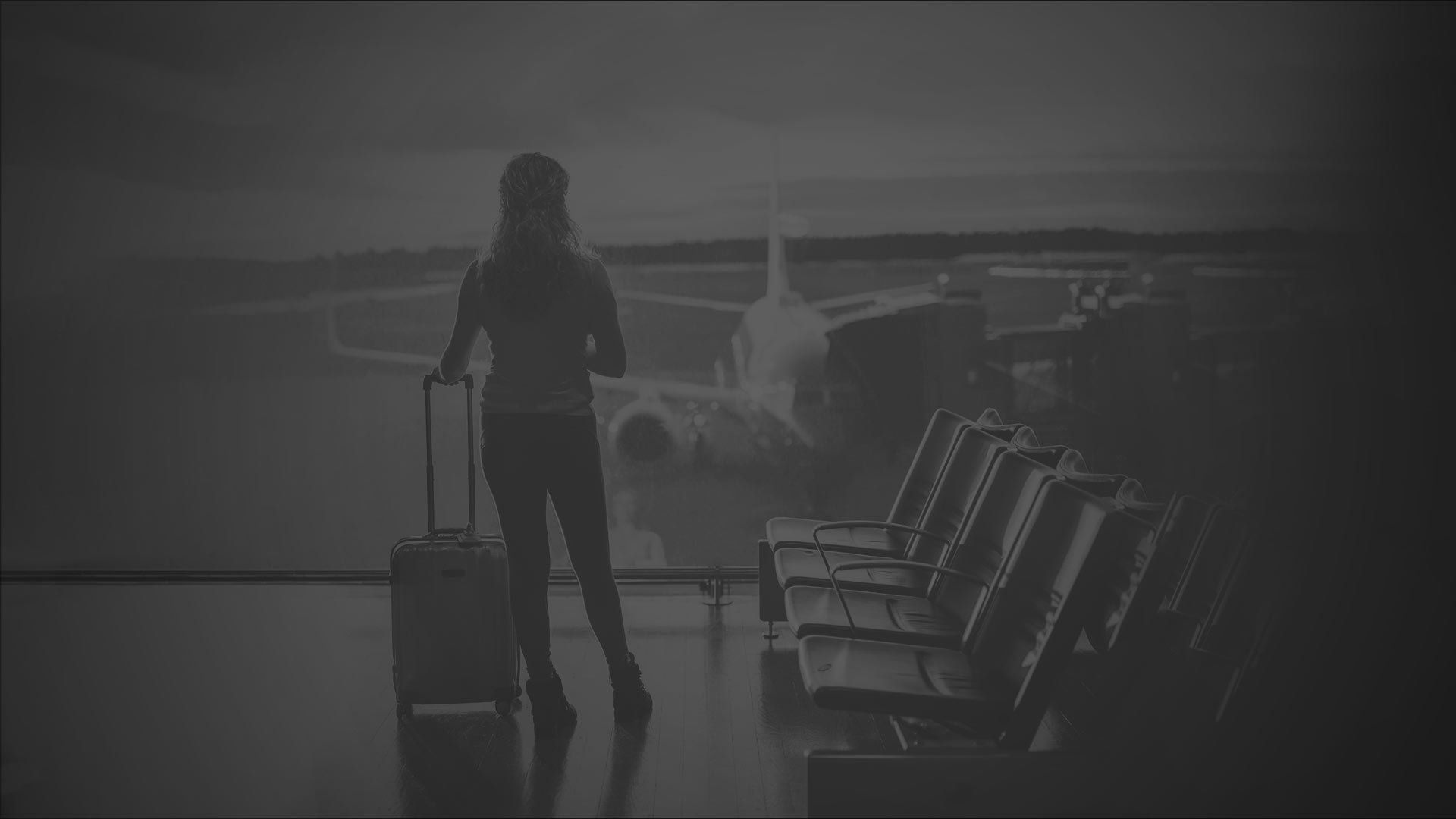 A woman stands in an airport terminal, holding luggage and looking around.