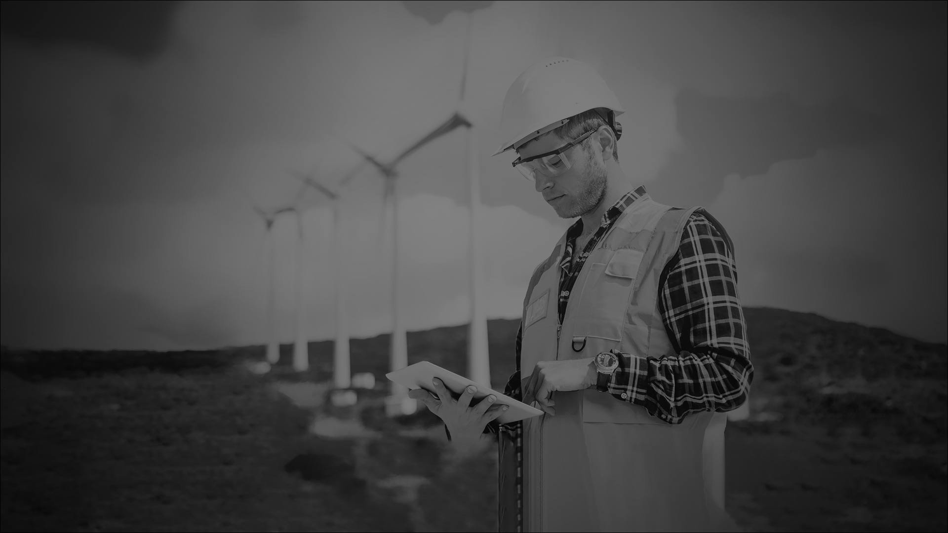 A worker in safety vest interacts with a tablet, with wind turbines in the background.