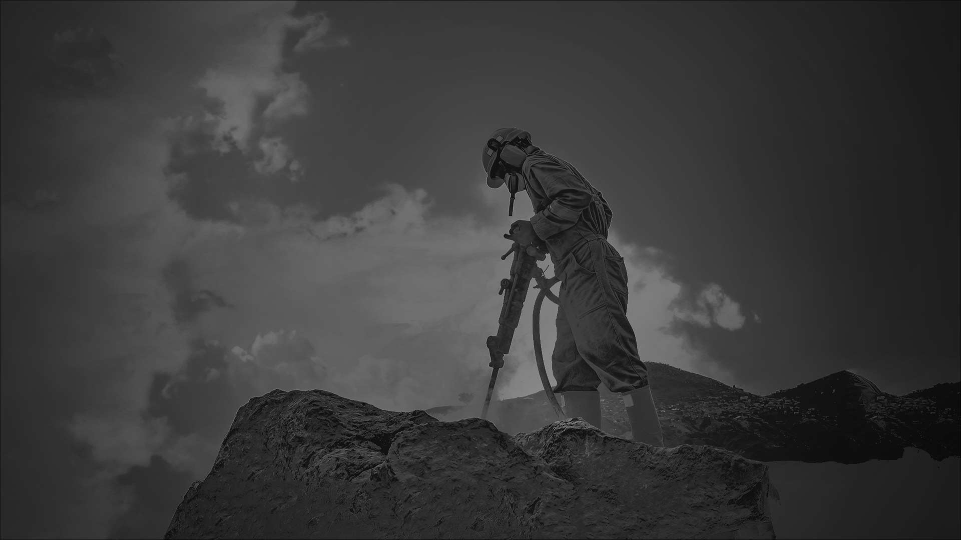 A worker wearing protective gear uses industrial equipment to drill into a large rock outdoors.